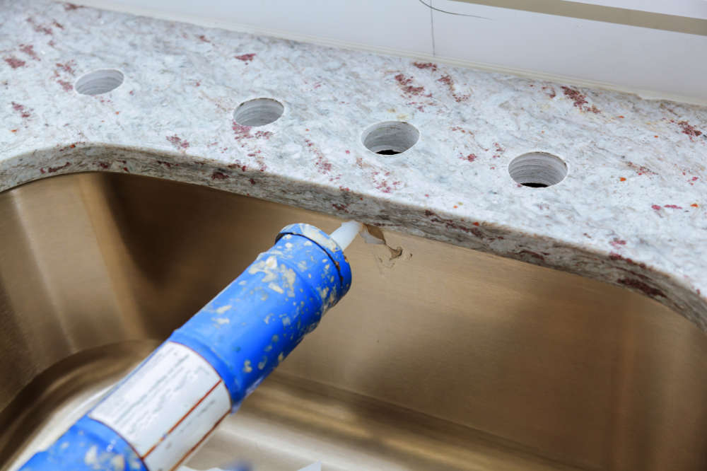 close up of new granite countertop installation and a person putting caulking around the existing sink under-mount